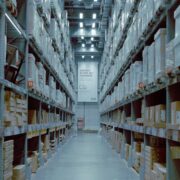 Long warehouse aisle between tall metal shelving units stacked with cardboard cartons under overhead lights
