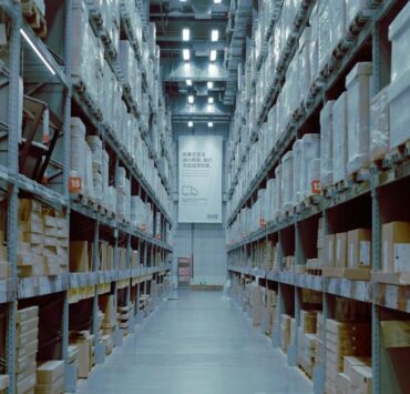 Long warehouse aisle between tall metal shelving units stacked with cardboard cartons under overhead lights