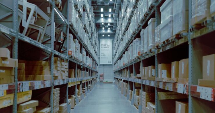 Long warehouse aisle between tall metal shelving units stacked with cardboard cartons under overhead lights